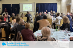 A picture of a room of people watching a horse race on a projection screen. at the bottom is a white band with Bury North and South Conservatives in Light Blue