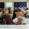 A picture of a room of people watching a horse race on a projection screen. at the bottom is a white band with Bury North and South Conservatives in Light Blue